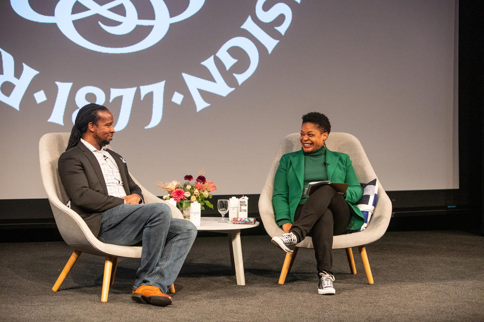 A smiling Black man and woman sit on a stage. Between them is a table with flowers and boxes of water. The RISD seal is projected behind them.