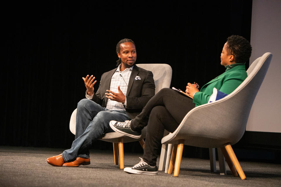 A Black man and woman sit on a stage. The man gestures and speaks as the woman looks on.