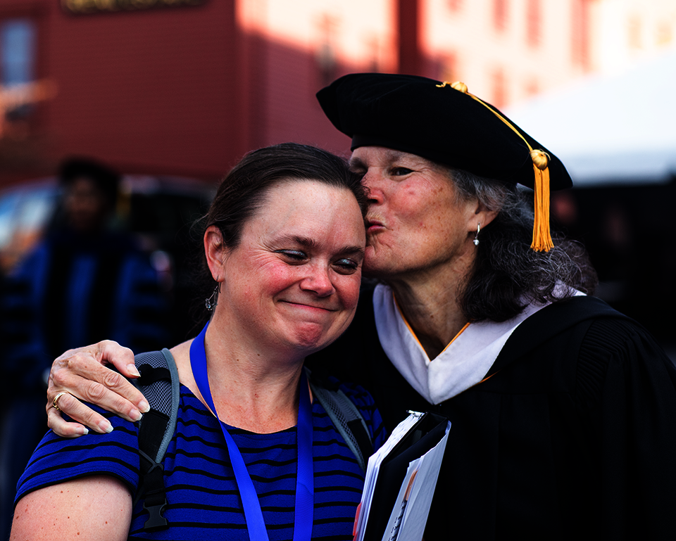 A white woman in doctoral robes embraces a smiling white woman in blue
