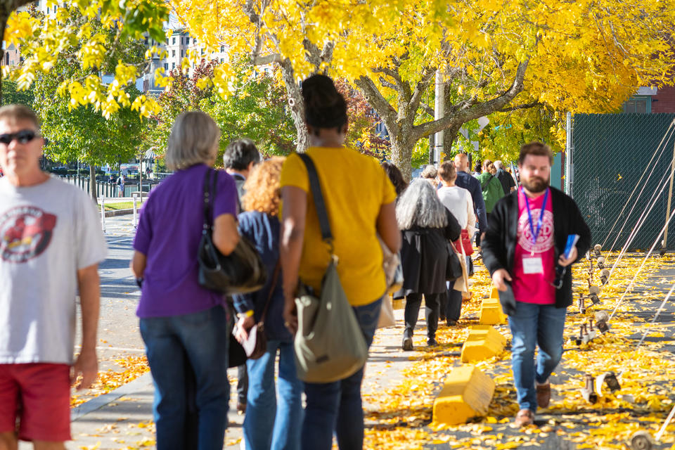 Crowds of people walk down a tree-lined sidewalk. The trees are full of bright yellow leaves.