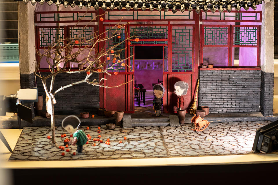Small diorama set of a scene outside a house with Asian style architecture. In the foreground, and young girl figure picks up persimmons. 