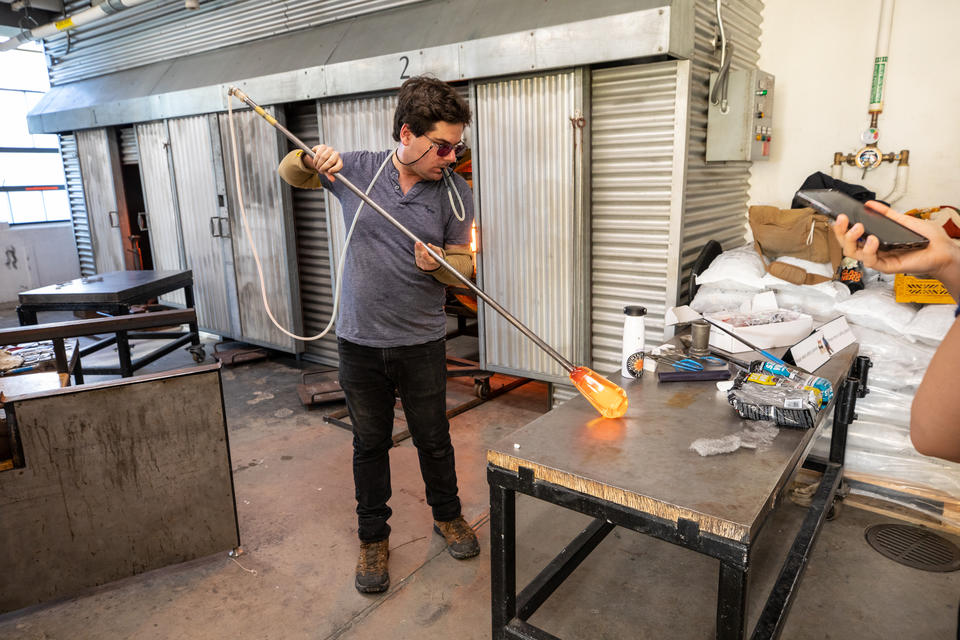 A man demonstrates blowing glass. He holds a pipe with molten glass on the end, rolling it on a table as he blows into a tube connected to the pipe.