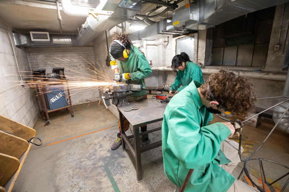 Three students work in a metals shop wearing green protective smocks. Sparks fly as one student cuts through metal.