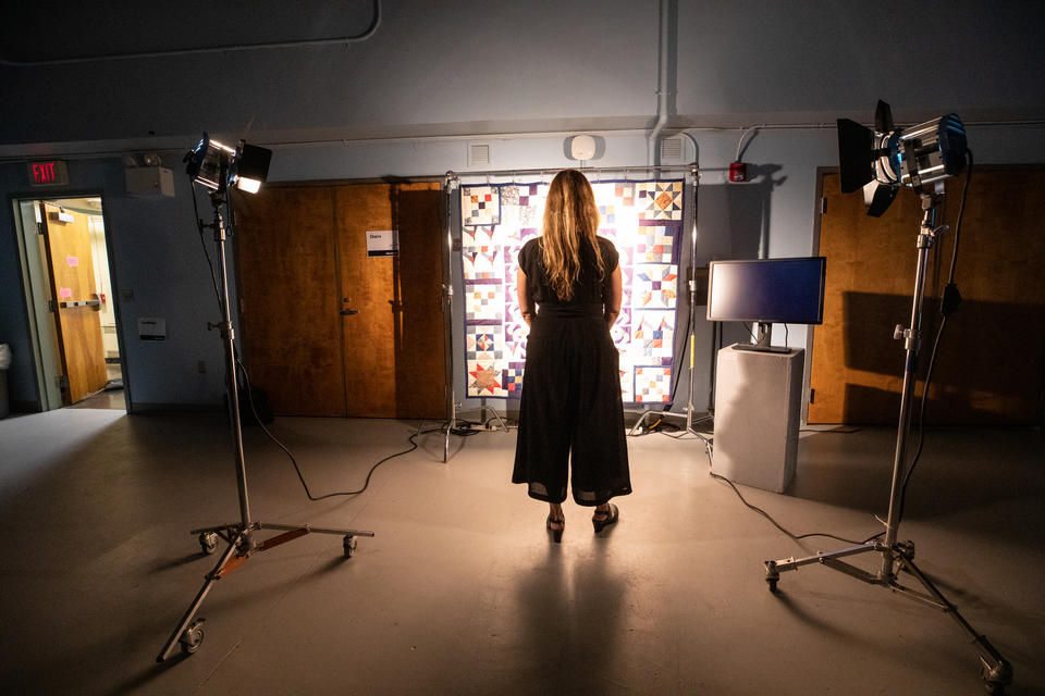 A woman stands in a classroom, in front of a suspended quilt lit by two powerful fresnel lights with barn doors.