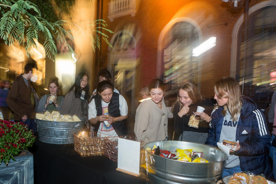 Nighttime photo of several young women looking at a sign in front of a table of snacks.