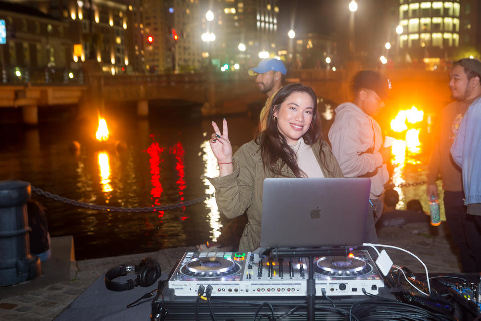 At night, a smiling young woman stands in front of DJ equipment, raising her hand in a peace symbol. She has long green nails. In the background, braziers burn in the Providence river.
