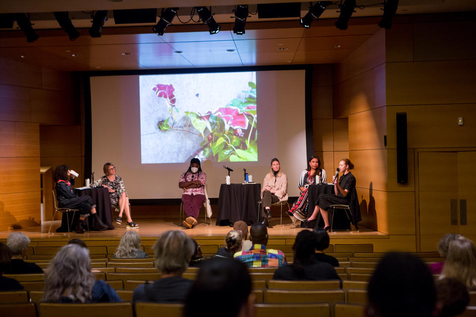 Four panelists and two moderators sit on stage at a wood-paneled auditorium. An image of a flowering vine is projected behind them.