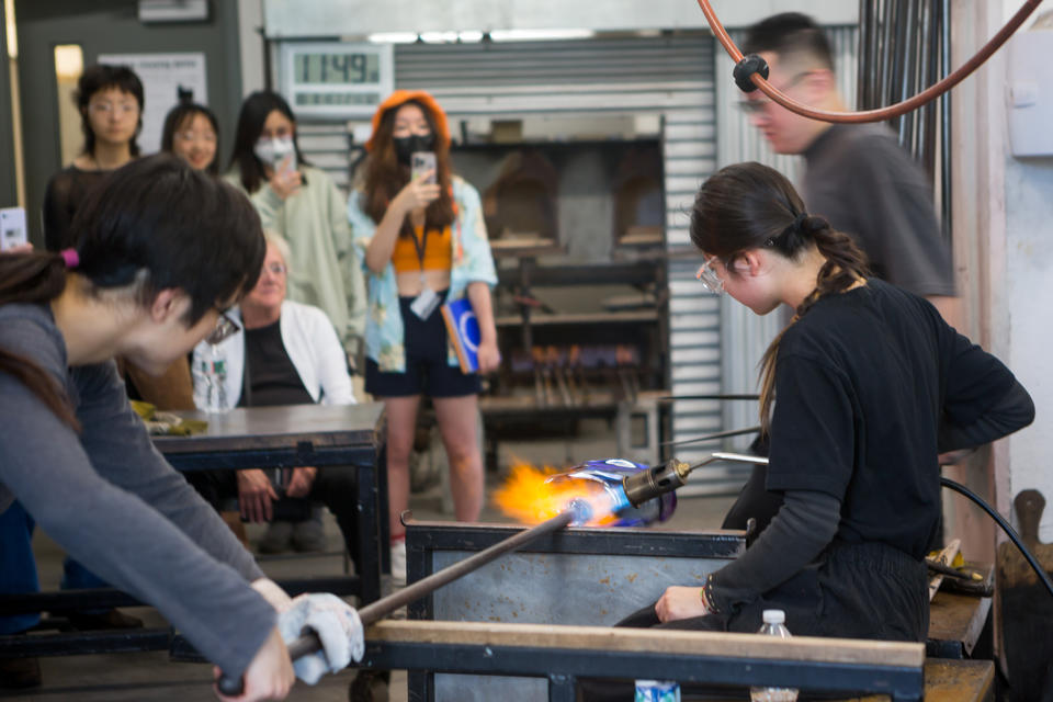 Two students demonstrate blowing glass, using a torch to heat glass on a long pipe,  while others look on, recording with their cellphones.