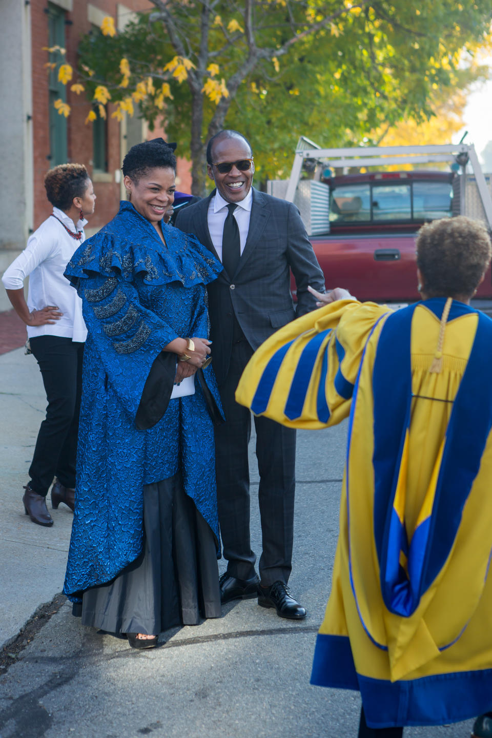 A Black woman in blue ceremonial robes and a Black man in a suit greet a woman in yellow and blue doctoral robes