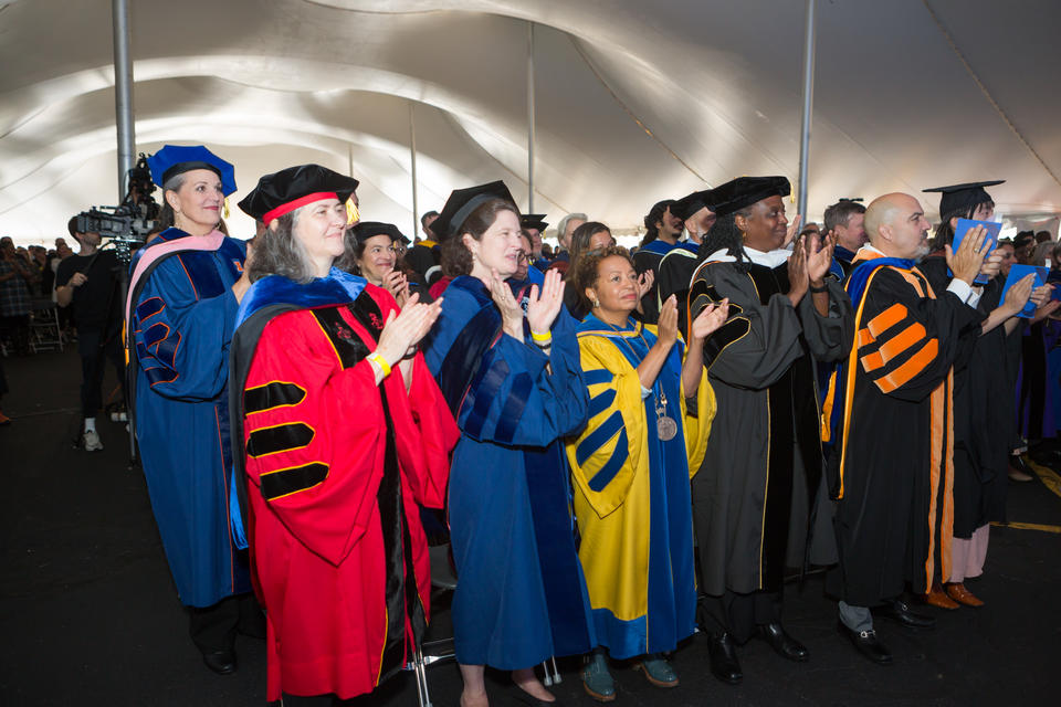 A group of adults in doctoral robes of various colors stand and clap in a tent
