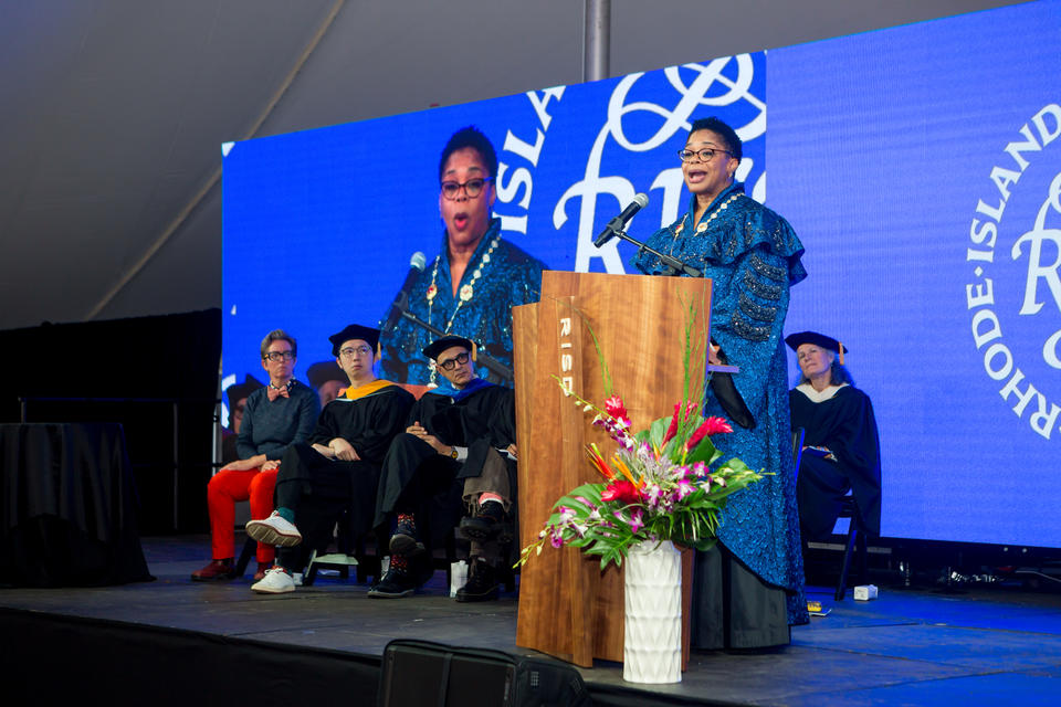 A Black woman in blue ceremonial robes speaks at a podium, on a stage where a group of onlookers sit. Her image is projected behind them.