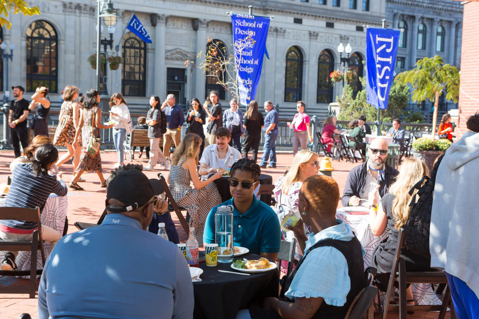 On a sunny Fall day, groups sit and eat at small round tables. In the background, people walk along the Providence Riverwalk. Blue RISD banners hang from light poles.