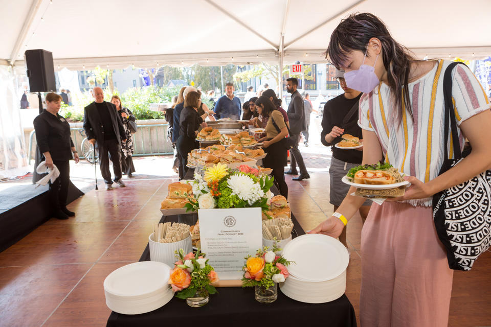 Buffet line outdoors under a tent. A table is piled with sandwiches, plates, and flowers. A young AAPI person picks up a plate in the foreground, while a small group examines food offerings in the background.