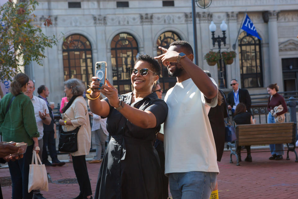 Outside on a brick walkway a Black woman in sunglasses takes a selfie with a young black man in a white tee shirt.