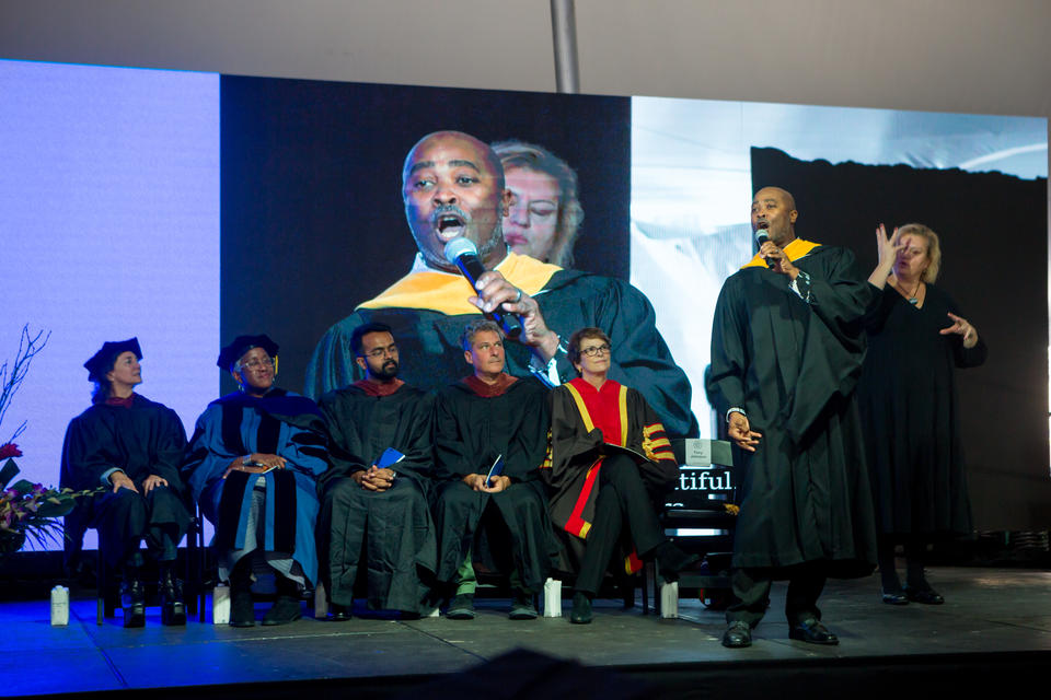 A black man in masters robes sings on a stage, with five professors seated, looking on. A woman stands behind him, interpreting in sign language.