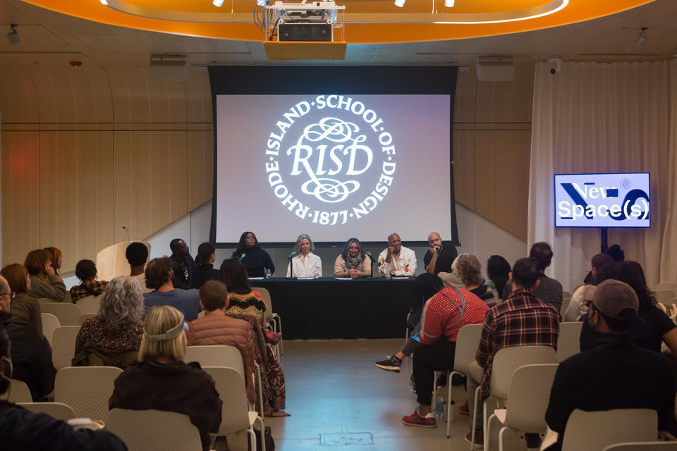 Five panelists and one moderator sit at a table in front of a small seated crowd. The RISD seal is projected behind them.