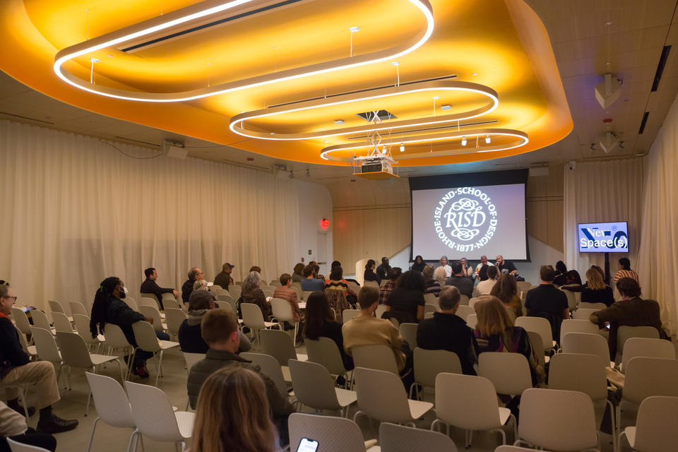 Five panelists and one moderator sit at a table with the RISD seal projected behind them. Large circular light fixtures hang above them.