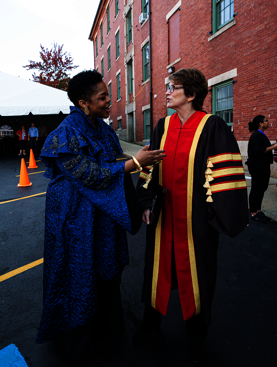 A Black woman in blue ceremonial robes engages in cheerful conversation with a white woman in doctoral robes with red and golds stripes.