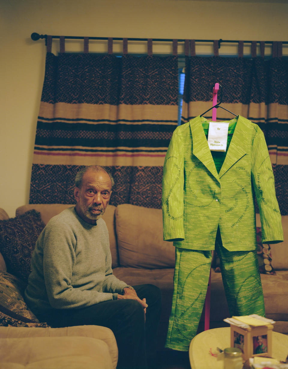 Man sitting in his living room on a couch with a green-colored suit hanging on a clothing rack to his right.