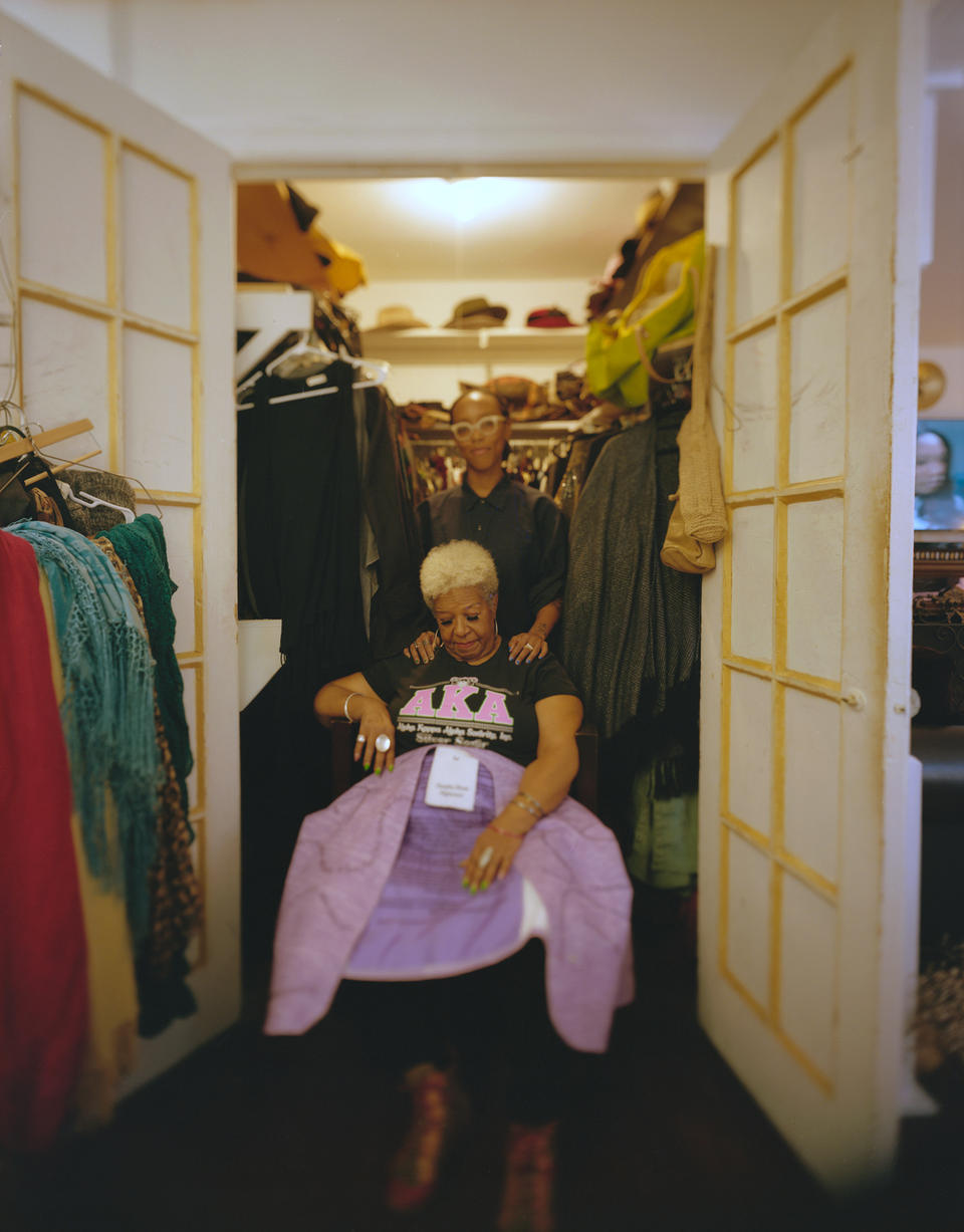Woman sitting on a chair at the entrance of a double-door closet. She is holding a purple dress and looking down at it upon her lap. Behind her stands a woman smiling and looking at the camera.