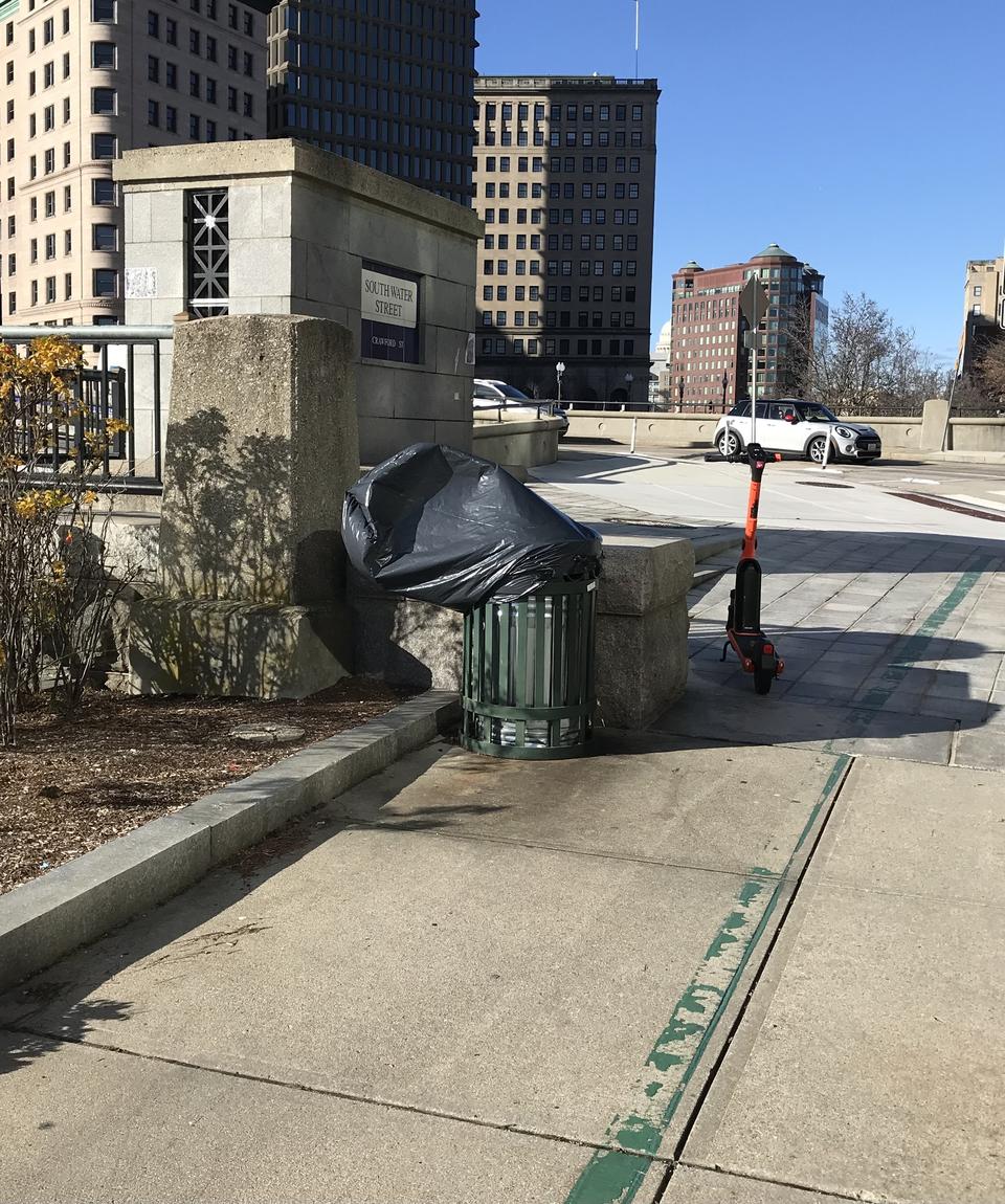 A trash can along the Providence River with the bag blowing around above the can in the wind.
