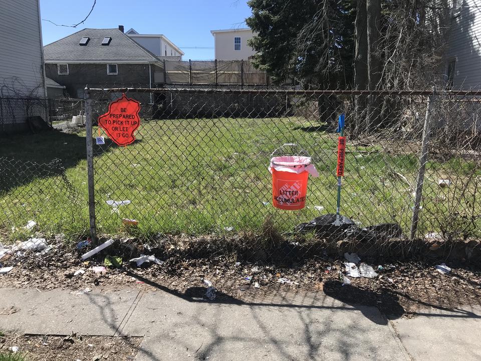 An orange sign, orange bucket, and trash grabber attached to a fence where litter has accumulated.