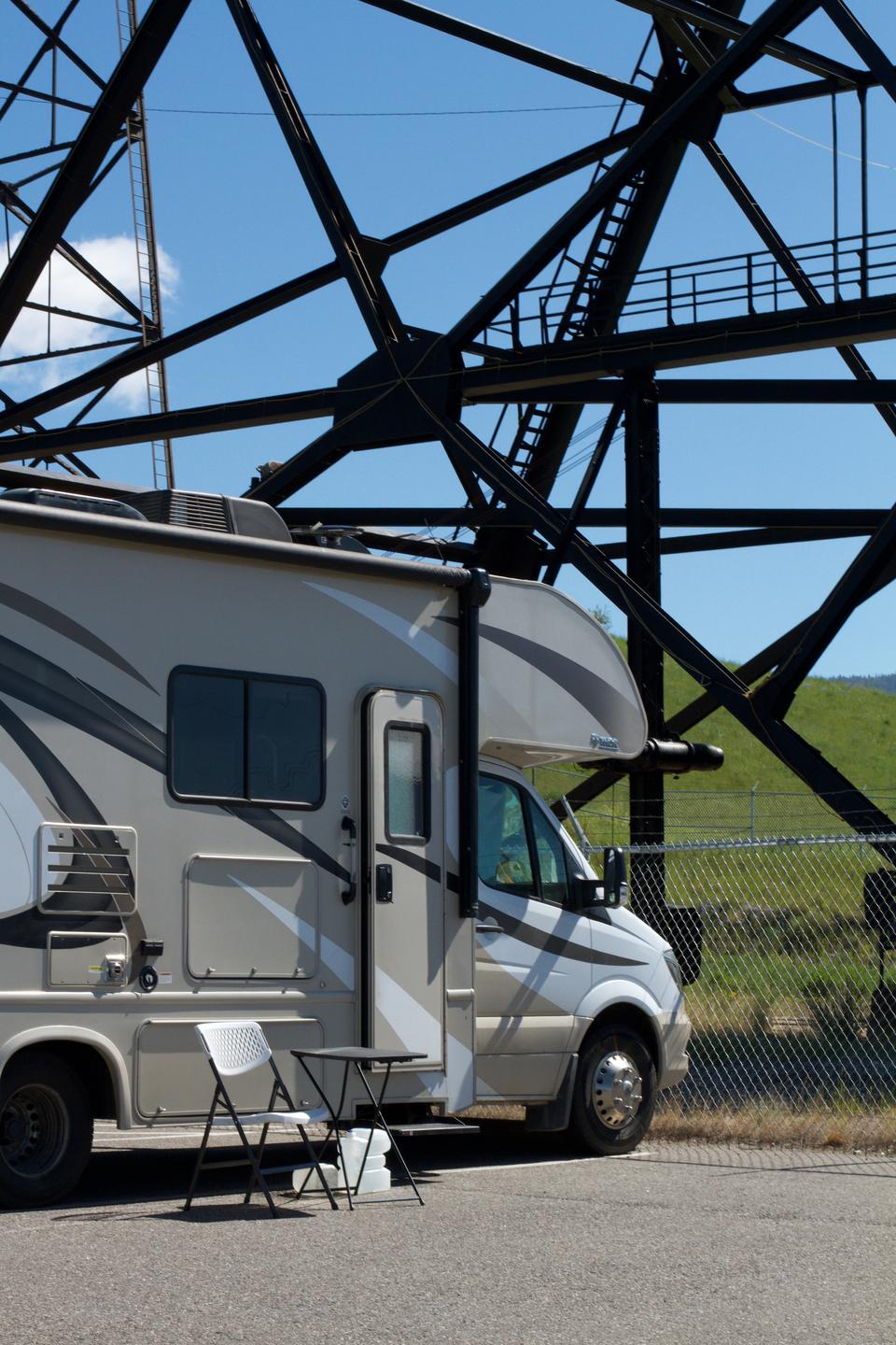 A camper van parked in front of the Mountain Con headframe. The swishy pattern of the camper's exterior seems to mirror the steel that criss-crosses the blue sky. The door of the camper is slightly ajar, and a camping chair and table sit on the parking lot outside the vehicle.. 