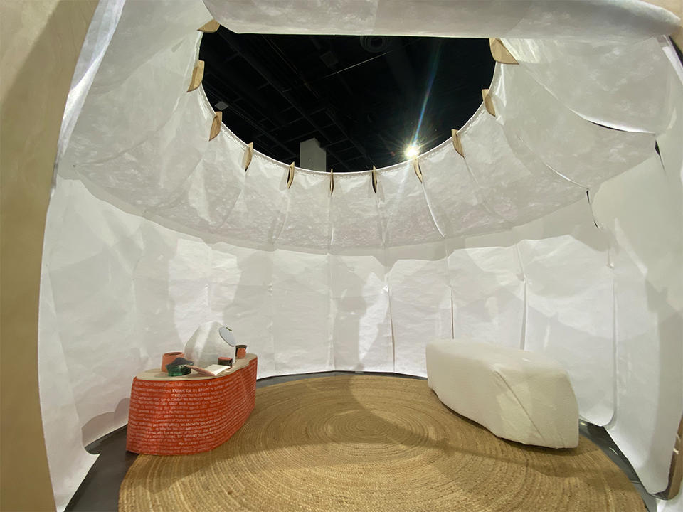 The interior of a white stand-alone structure houses an orange table with a white ceramic speaker, dried herbs, and cards.