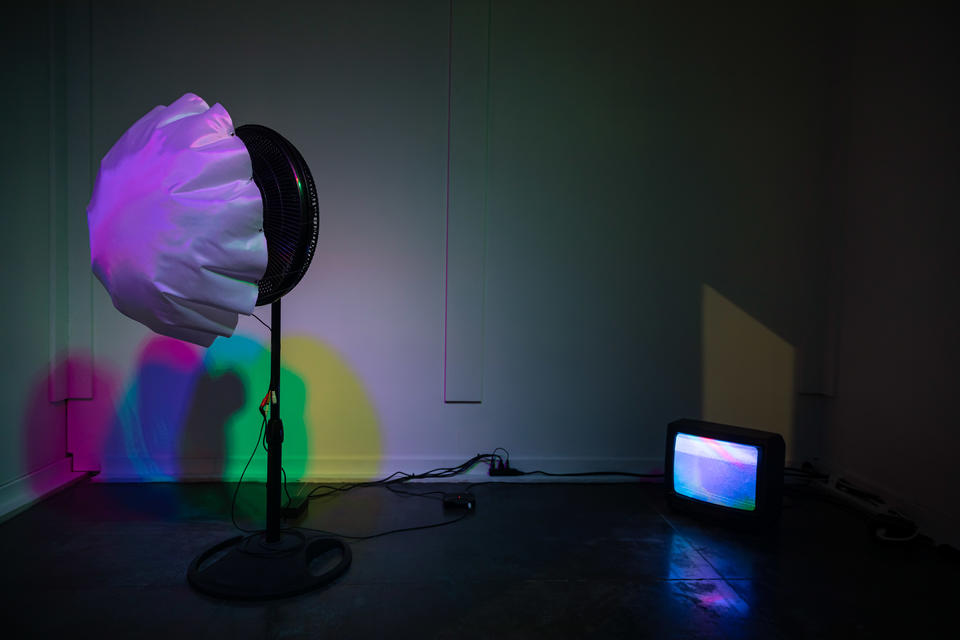 White fabric is attached to a fan and being blown up under colorful light.