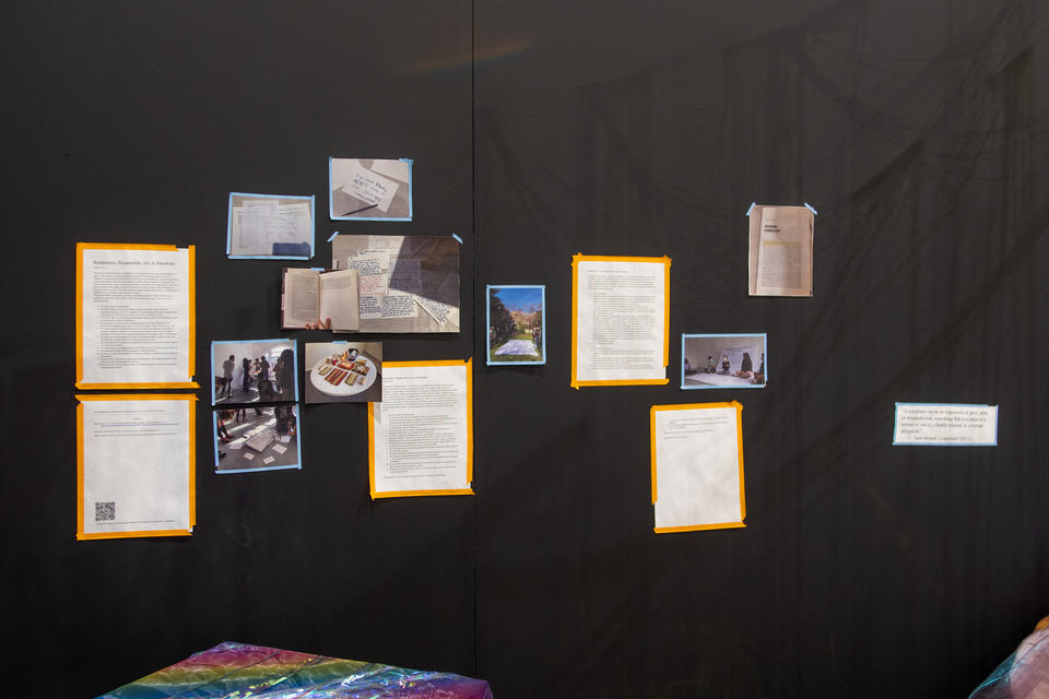 books, food, and drinks on white table; iridescent party streamers hanging from above like curtain; photos and writings on corner-shaped black walls