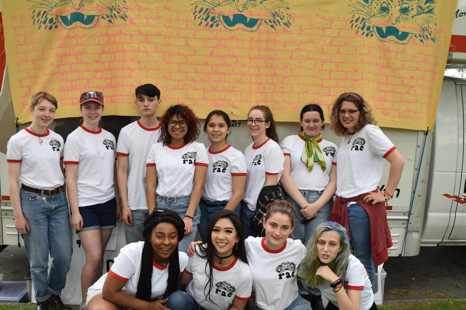 Twelve teenagers in two rows all wearing a white t-shirts with a maroon collar in front of beige fabric banner with blue monsters. 