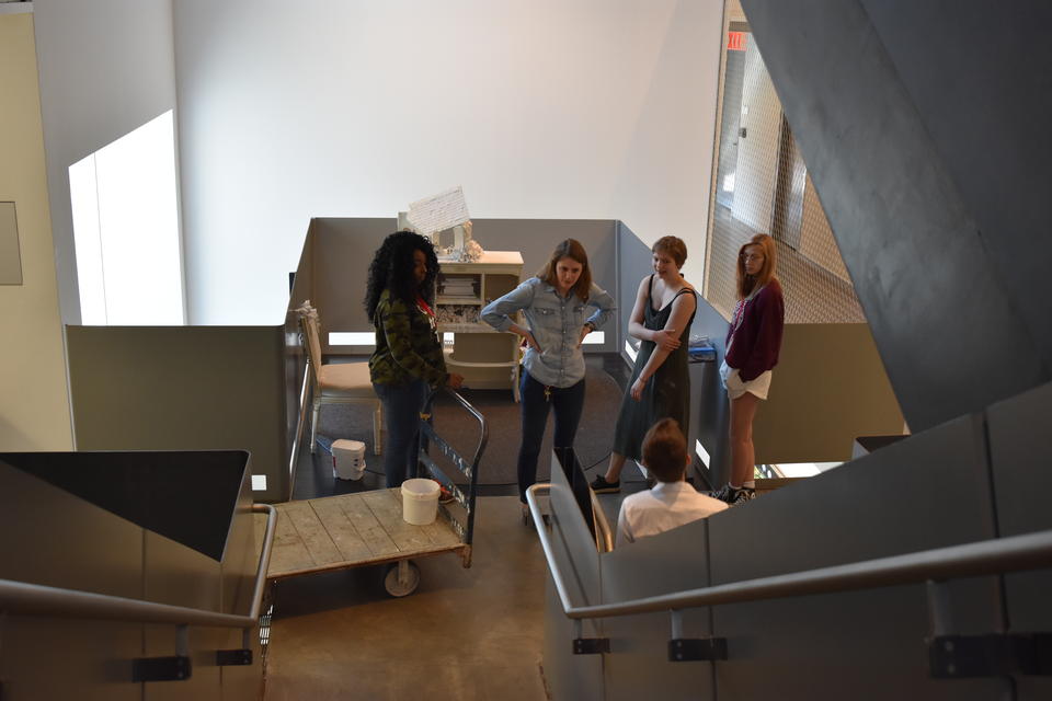 Camera angled from the top of the stair looking down a staircase at five people talking  with some art installation equipment nearby.