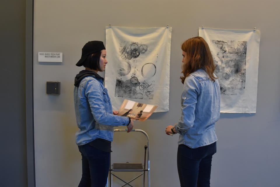 Two people talking with step-stool behind them but in front of piece of white fabric with a black ink drawing on it