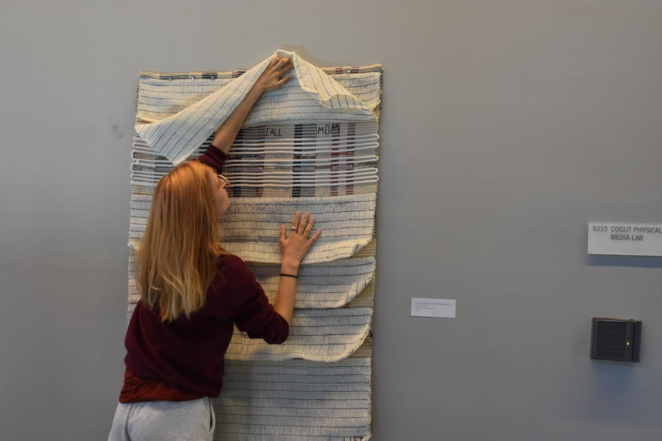 Student straightening a wall piece that has layers of fabric one of which is being lifted to reveal small letters that spell Call Mom.