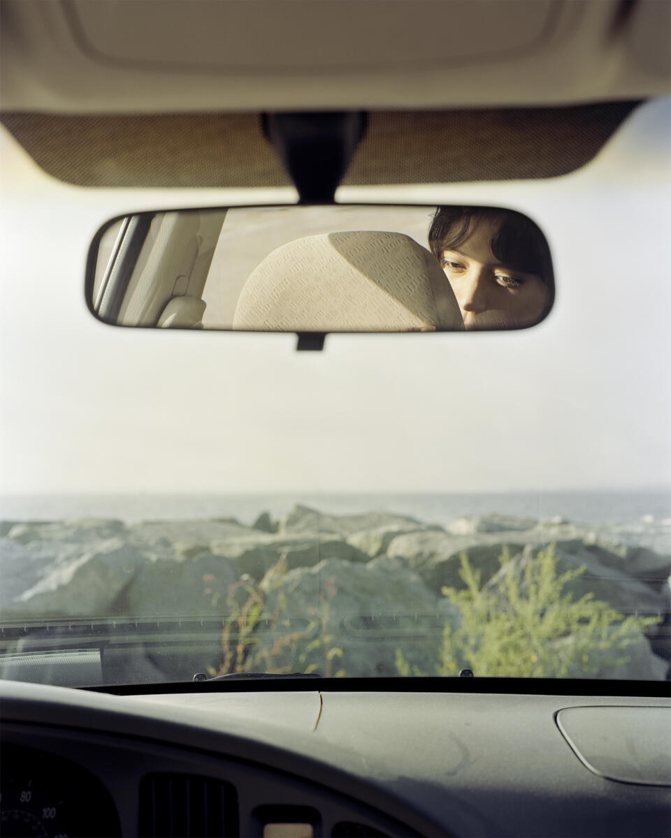 Photograph from inside of car looking out through the windshield to a rocky beach. The dashboard sits at the bottom of the image. There is a rearview mirror with a reflection of person face.