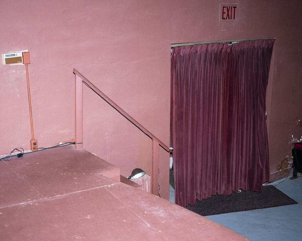 Photograph of a pink wall and stage. On the floor sits a doorway covered by a fushia velvet curtain. Behind the curtain an obscured person. Exit sign sits above the curtain  