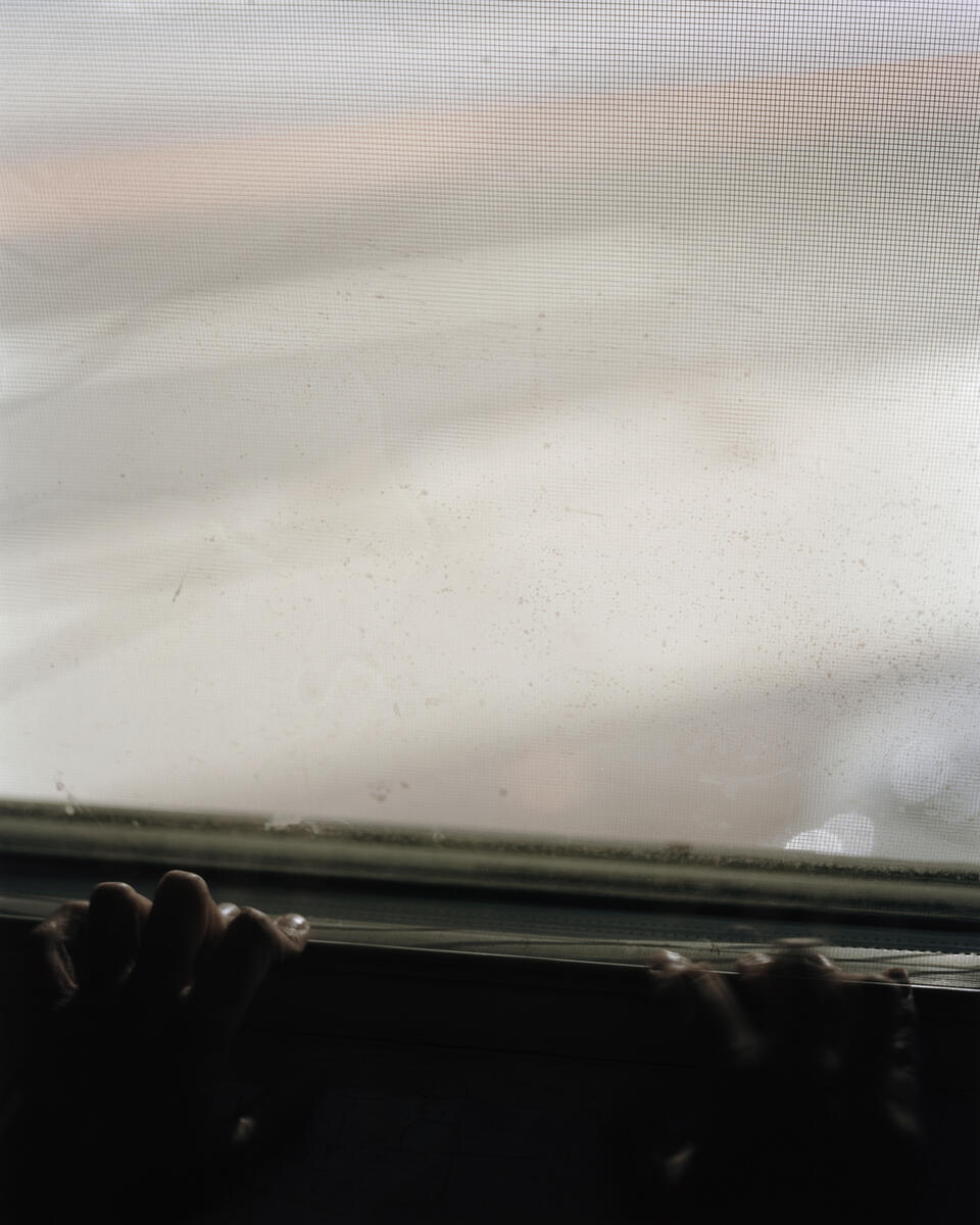 A long exposer photograph of a hands resting a on a white windowsill at night. The hands are mostly in shadow. Through the window is the gray street with long obscured car lights. 