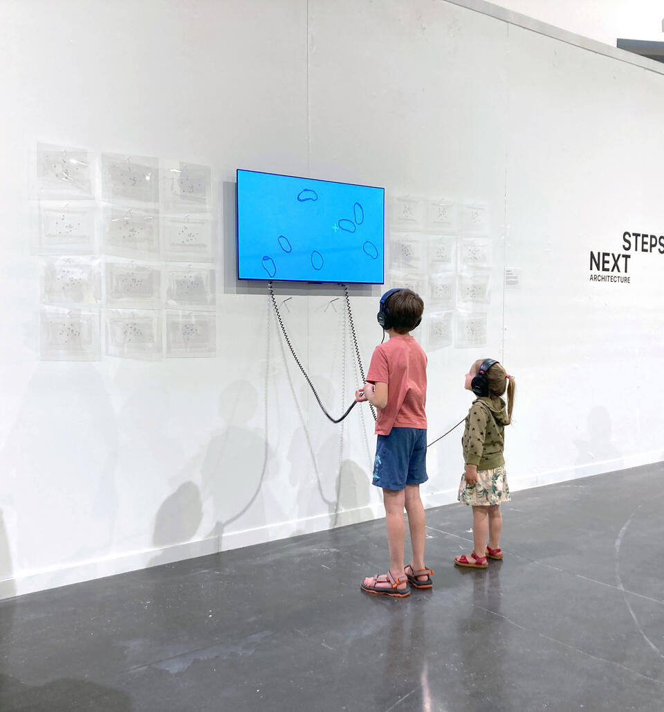 Two small children, both wearing headphones, gaze upward to watch a film installation. Shapes on the screen move against a backdrop of blue sky.