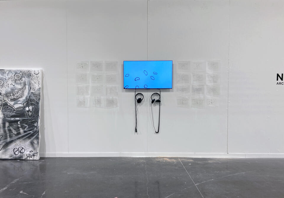 A TV is mounted on a white wall. On screen, shapes move against the background of a blue sky. Many small, transparent sheets hang neatly around the TV, two pairs of headphones hang beneath.
