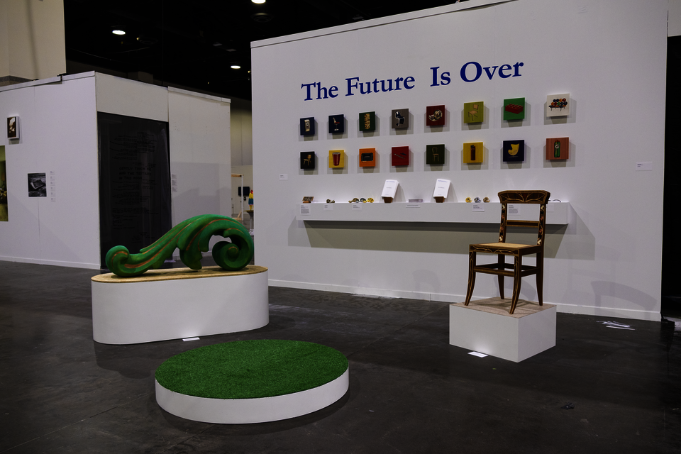 Large green acanthus ornament and a chair in front of a wall with 16 panels depicting chairs and ubiquitious products, a shelf with jewelry-scale objects, and the words "The Future Is Over" in blue letters 