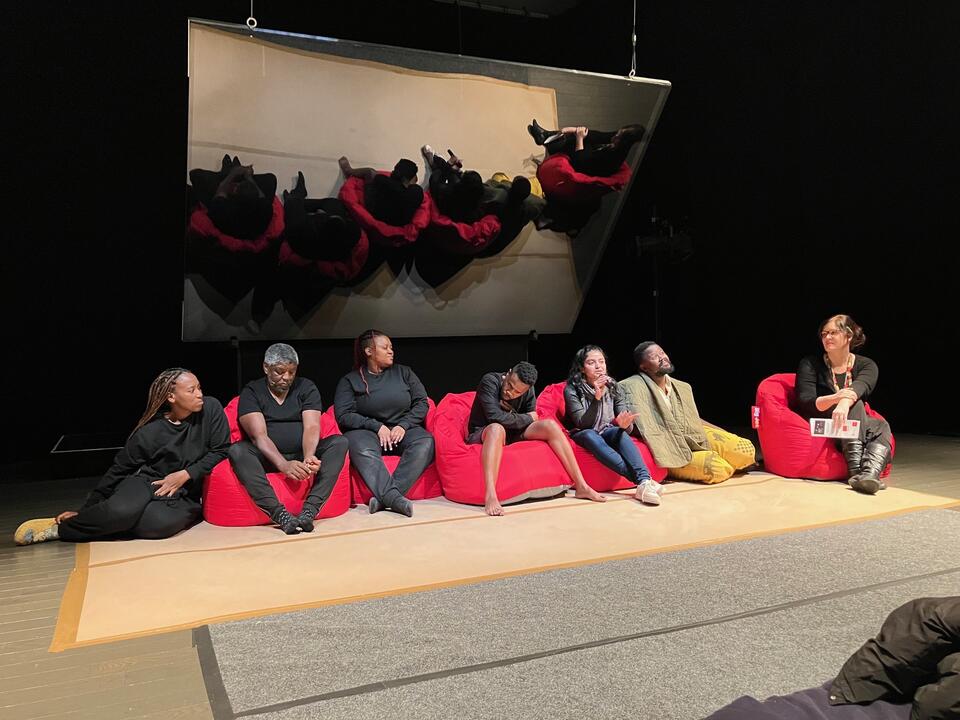 Individuals seated on red bean bags in front of the Pepper's Ghost Theatre Apparatus. Collaborators from William Kentridge's Centre for the Less good idea. Collaboraters are South African. Deanne Fernandes is present as well. 