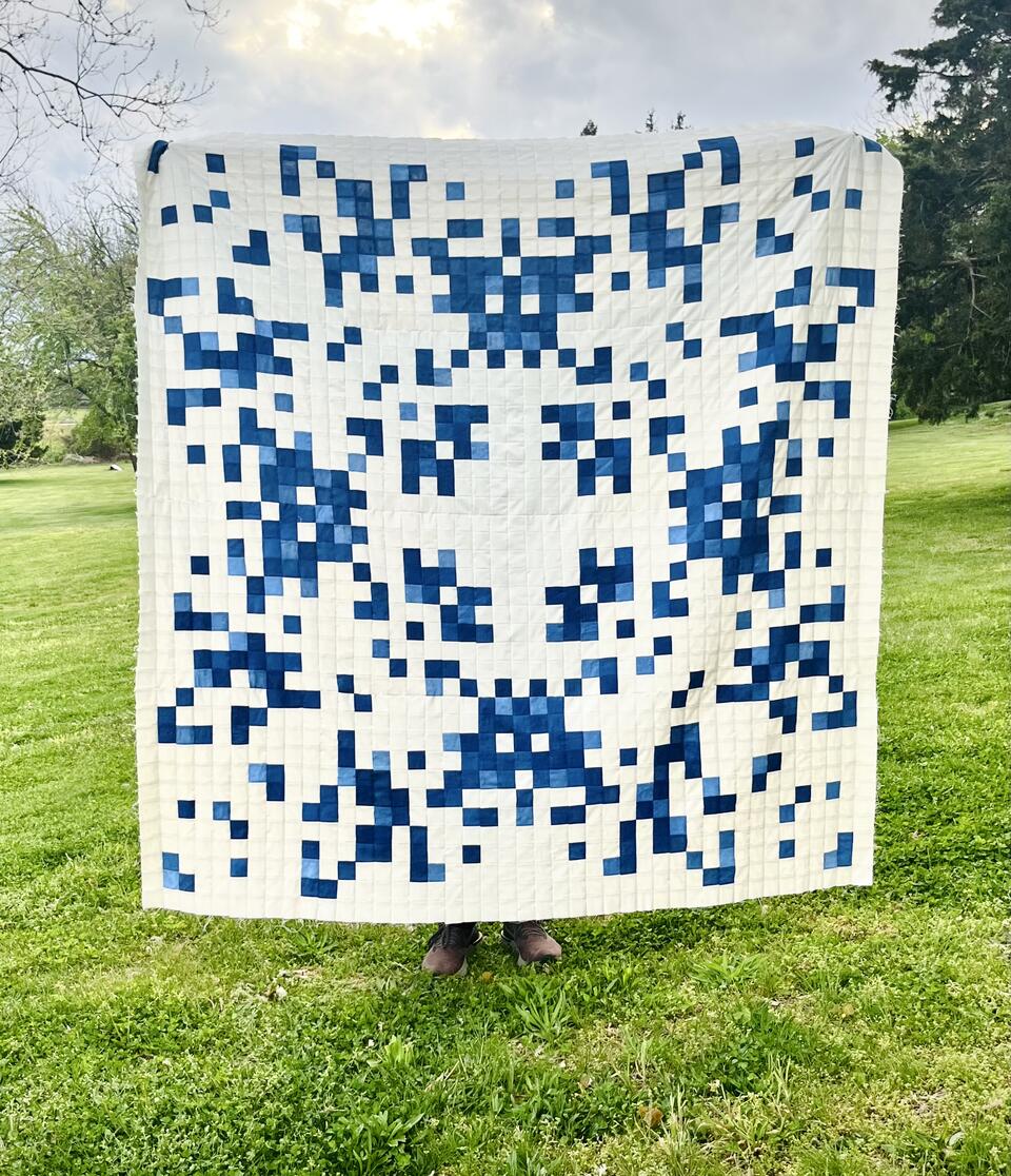 A large blue and white quilt is held up outside in a field of green grass at golden hour.