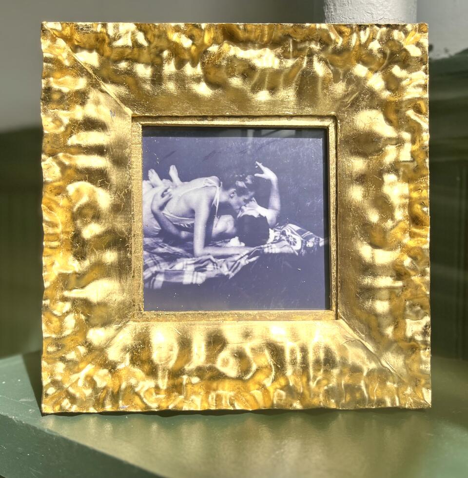 A golden frame holds a black-and-white vintage photo of two people kissing on a blanket in the park. 