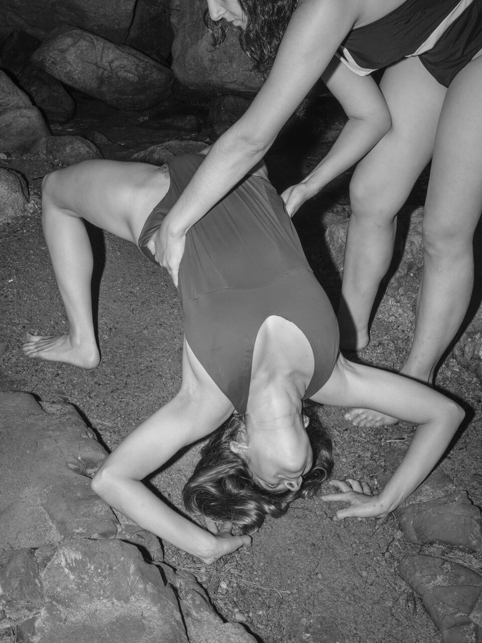 Black and white photograph of two people on the rocks of a beach, one person holding the other while she tries to make a complicated body posture