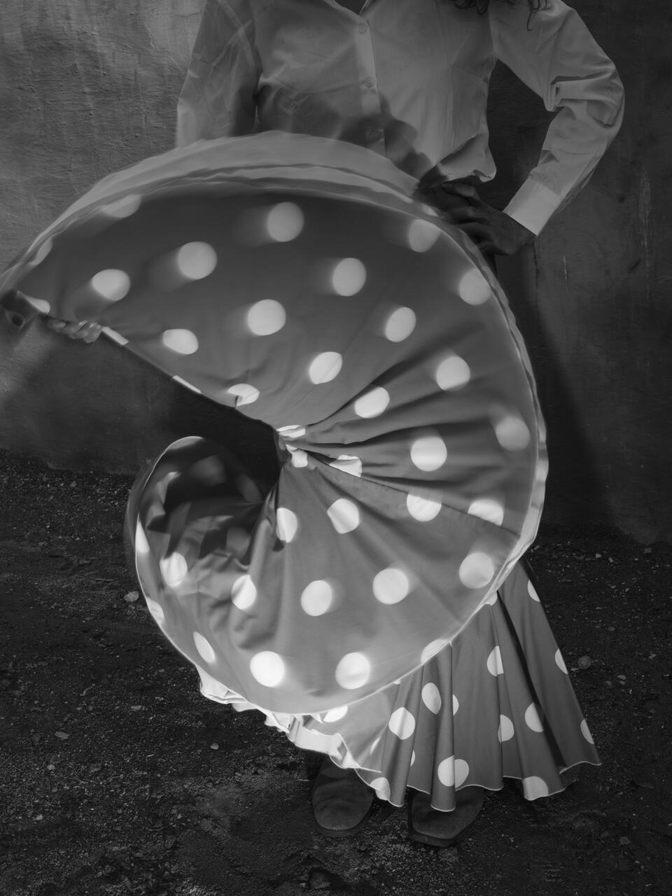 Black and white photograph of a person with a skirt full of white dots, which they are undulating with one hand for the picture 