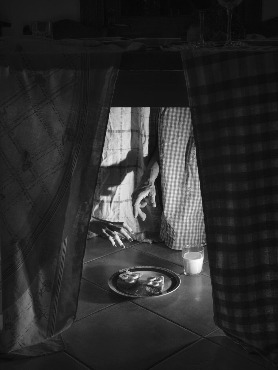 Black and white photograph of the bottom of a table, with 4 hands almost grabbing a plate of bread with banana and a glass of milk