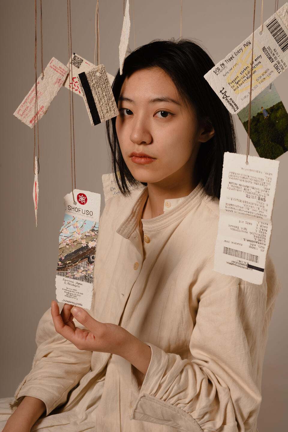 A girl wearing a cream dress surrounded by suspended tickets painted on paper mache. Her one hand is touching one ticket, eyes looking at the camera. 