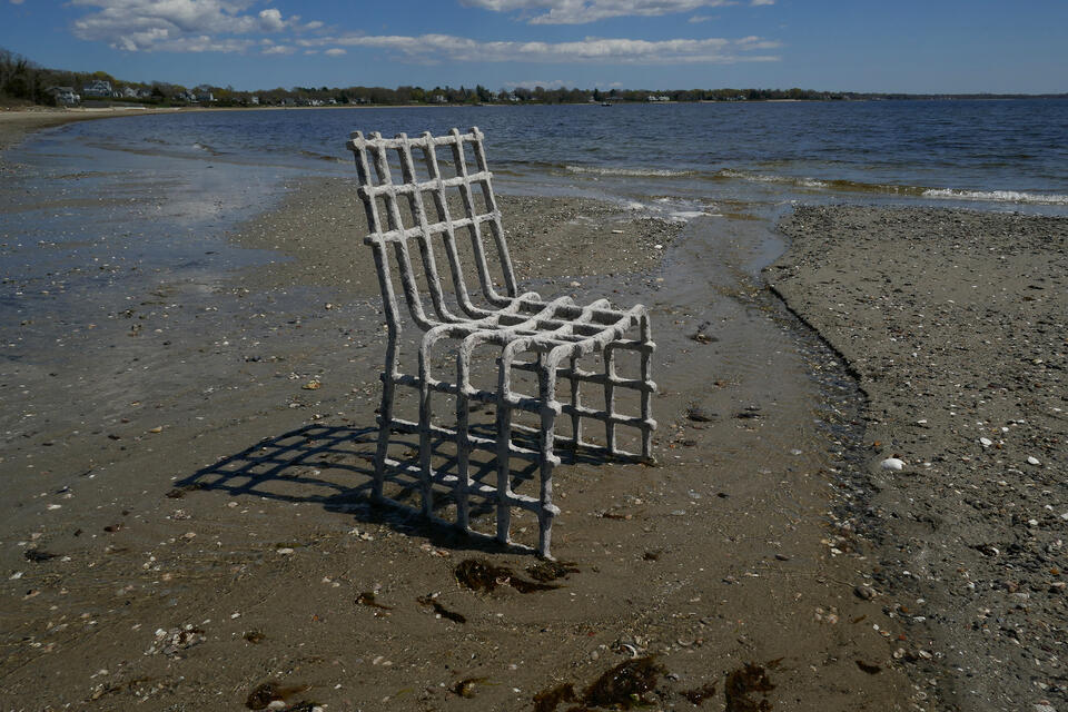 A gray and blue oyster and mussel biomaterial chair on a sandy, rocky beach by the water's edge. Houses and trees are visible across the water under a partly cloudy sky.