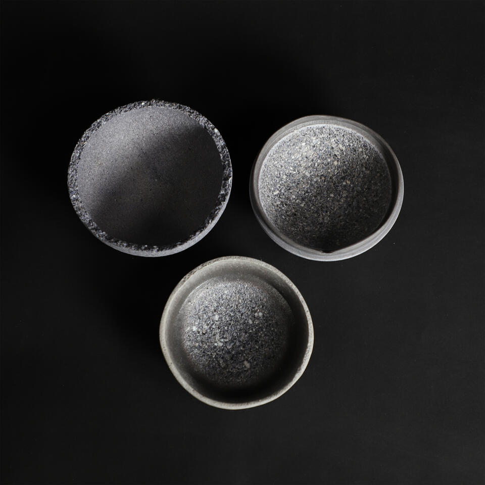Three shallow, round bowls made from mussel shells, arranged in descending size on a black background. The bowls have textured, dark blue exterior surfaces and smooth, light gray and blue interior finishes.