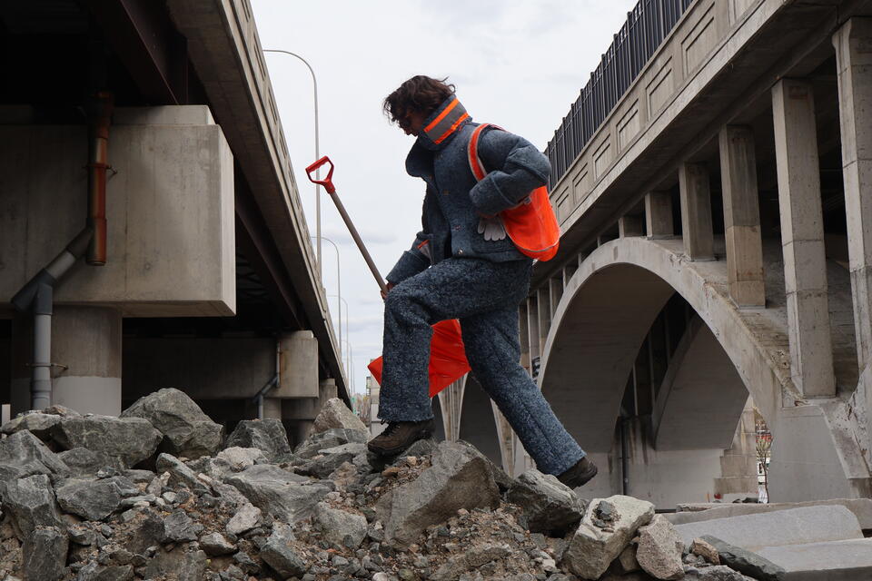 Person wears Recycled Denim Suit climbing on a pile of construction rubble holding a shovel
